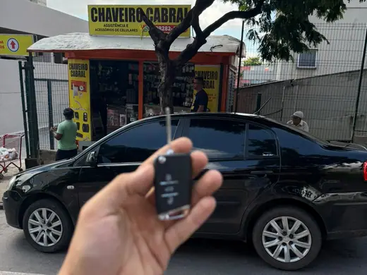 Chave canivete na mão, com um sedã preto e o quiosque do Chaveiro Cabral ao fundo