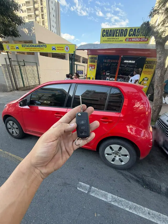 Mão segurando uma chave canivete pronta, com um VW up! vermelho e o quiosque amarelo do Chaveiro Cabral ao fundo