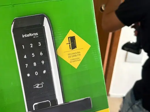 Caixa de uma fechadura digital Intelbras com teclado numérico e, ao lado, o instalador com a furadeira, durante uma instalação